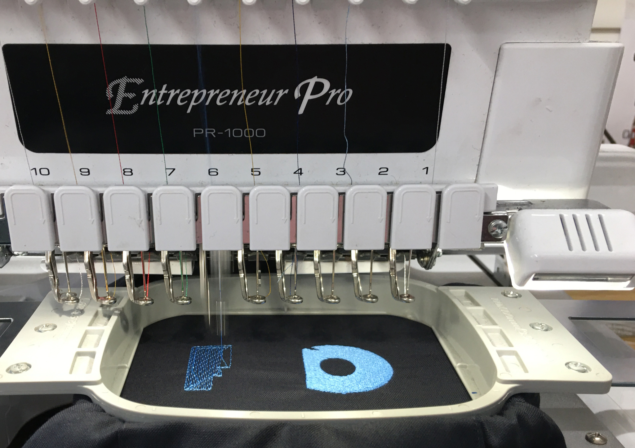 Embroidery machine with 10 different thread colours in motion stitching a blue logo on a black shirt.